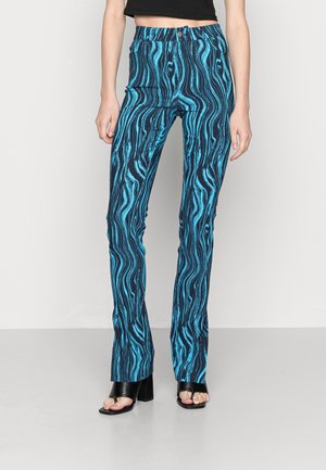 Femme portant un pantalon taille haute à motif ondulé bleu et noir, associé à des sandales à talons blocs noires à bouts ouverts, debout sur un fond uni.