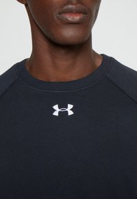 Primo piano di una persona che indossa una maglietta blu scuro con il logo bianco di Under Armour centrato sotto il colletto.