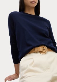 Navyblauer Pullover aus weichem Stoff, kombiniert mit cremefarbenen Hosen und einem hellbraunen Leder Gürtel mit einer runden, geflochtenen Schnalle.
