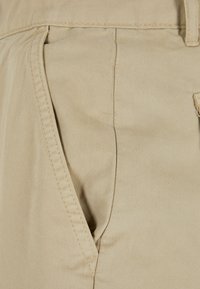 Tela de algodón beige con una textura suave, que presenta bolsillos laterales y un diseño de frente plano. Detalles de costura sutil a lo largo de los bordes.