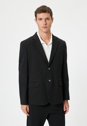 Zwarte blazer met een enkelvoudige sluiting, twee voorzakken en zwarte knopen. Draag het over een witte blouse met kraag, met een gladde textuur.
