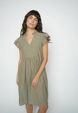 Žena s dlhými tmavými vlnitými vlasmi, oblečená v voľných, po kolená dlhých olivovo zelených šatách s volánikovými krátkymi rukávmi a výstrihom do V, stojaca pri bielej stene.