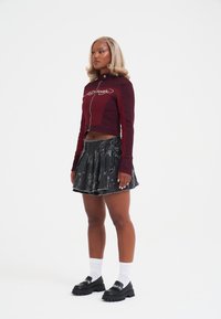 Kobieta z blond włosami stoi, ubrana w bordową kurtkę na zamek, czarną mini spódniczkę z nadrukiem, białe skarpetki oraz czarne masywne buty.