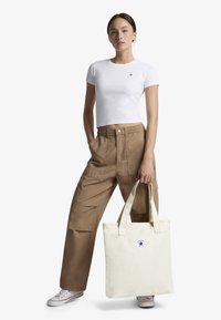 Balta trumpa marškinėliai, rudi krovinių tipo kelnės su kišenėmis ir šviesios spalvos didelė rankinė su apvaliu logotipu; įvaizdį baigia Converse sportbačiai.