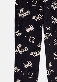 Černé fleecové kalhoty s bílými grafickými vzory, včetně obrysů koček a frází jako "Good Vibes" a "Love", na texturovaném pozadí.