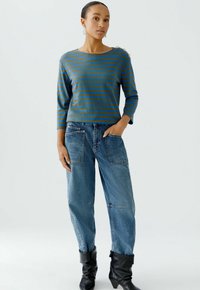 Gestreiftes, blaues und grünes Langarmshirt mit Schulterknöpfen, kombiniert mit weit geschnittenen blauen Jeans aus Denim, die über Taschen und kontrastierende Nähte verfügen.