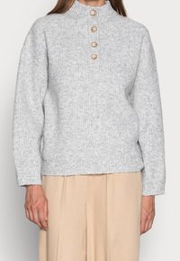 Hellgrauer Rippstrickpullover mit hohem Kragen und vier Holzknöpfen an der Vorderseite. Lockere Passform mit langen Ärmeln und geradem Saum.