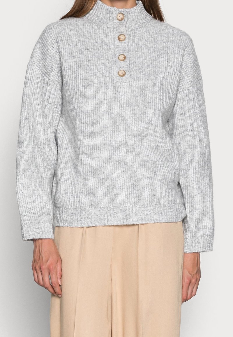 Hellgrauer Rippstrickpullover mit hohem Kragen und vier Holzknöpfen an der Vorderseite. Lockere Passform mit langen Ärmeln und geradem Saum.