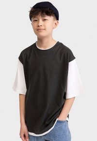 Tmavě šedé oversized tričko s bílou spodní vrstvou, krátké rukávy, kulatý výstřih a modré džíny. Stylizováno s černou čepicí.