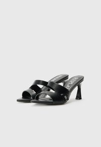 Sandales à talons noirs avec un bout ouvert et des brides croisées. Fabriquées en matériau lisse, elles présentent un petit accent décoratif sur le côté.
