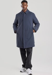 Navy blue, water-resistant coat with a collar, long sleeves, and side pockets. Worn over dark jeans and sneakers. Simple, streamlined design.