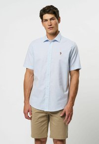 Lichtblauw kortavel shirt met knoopsluiting en een subtiel ruitpatroon, met een logo op de borst. Gecombineerd met beige shorts. Glad stoftextuur.