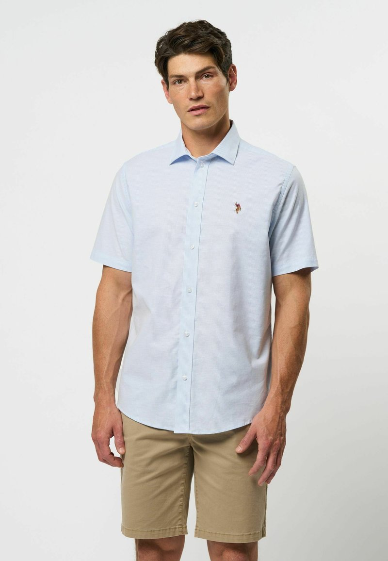 Lichtblauw kortavel shirt met knoopsluiting en een subtiel ruitpatroon, met een logo op de borst. Gecombineerd met beige shorts. Glad stoftextuur.