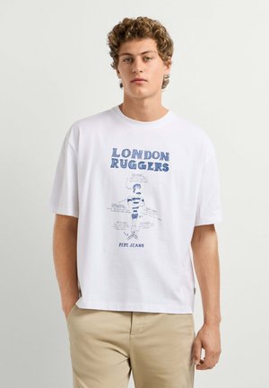Jeune homme portant un t-shirt blanc avec un graphisme "London Ruggers" et un pantalon beige, debout devant un fond clair uni.