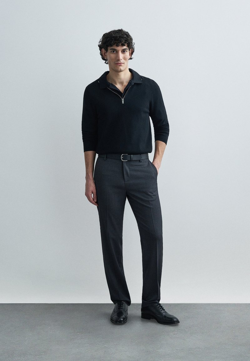 Black knitted zip sweater with a collar, paired with dark gray tailored trousers and black leather shoes. Model stands with hands in pockets.