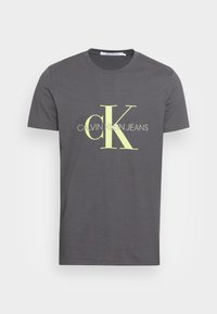 Šedé bavlněné tričko s výrazným, jasně žlutým logem "CK" a textem "CALVIN KLEIN JEANS" v čistém seřezaném písmu. Krátké rukávy.