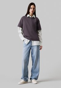Camiseta gris de gran tamaño superpuesta sobre una camisa de cuello verde claro, combinada con jeans de pierna ancha en azul desvaído y zapatillas color crema.