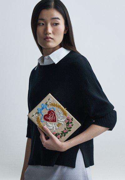 Bőr kézitáska hímzett virág- és galambdísszel, bézs alapon piros szíves részlettel és kék szalaggal. A modell fekete pulóvert visel.