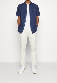 Camicia in denim blu scuro a maniche corte con bottoni sopra una t-shirt bianca a girocollo, abbinata a pantaloni beige a sigaretta e scarpe da tennis bianche con dettagli verdi.