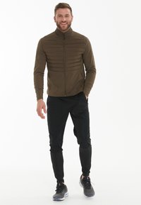 Olijfgroene zip-up jack met een horizontaal quiltpatroon, gecombineerd met zwarte tapered pants en grijze sportieve schoenen.