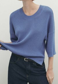 Pull en maille bleu avec un col rond et des manches trois-quarts, associé à un jean en denim foncé et une ceinture. Texture douce et coupe décontractée.