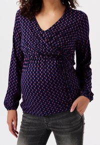 Schwangerschaftsbluse in Marineblau mit rosa und lila geometrischen Mustern. Sie verfügt über ein Wickeldesign, lange Ärmel und eine geraffte Taille mit Bindebändern.