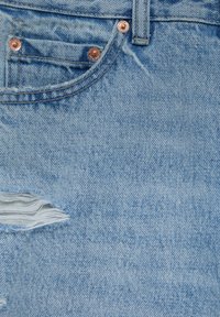 Tissu en denim bleu clair avec des rivets en cuivre, doté d'une petite poche avant et d'une déchirure horizontale révélant des fils blancs en dessous.