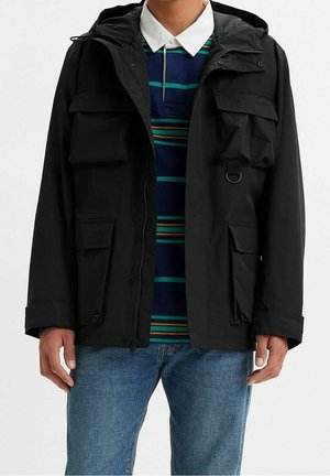 Mężczyzna w czarnej kurtce z kapturem nałożonej na niebieską koszulę w paski z białym kołnierzykiem oraz niebieskie dżinsy, stojący z rozluźnionymi ramionami.