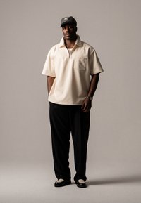 Pullover beige a maniche corte con collo a zip e tasca sul petto, abbinato a pantaloni neri e scarpe nere con un accento testurizzato.
