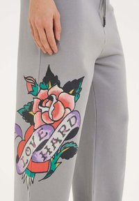 Grå sweatpants med en stor färgglad blommig grafik på vänster ben, med texten "LOVE HARD" i fet stil på ett band.