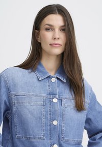 Lichtblauwe spijkerjas met een kraag, vier zakken en zilverkleurige knopen. Heeft een comfortabele pasvorm en een zachte textuur.