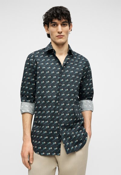 Νεαρός άνδρας που φοράει σκουρόχρωμο πουκάμισο με μικρά λουλουδάτα σχέδια και μπεζ παντελόνι, στέκεται μπροστά σε ένα απλό φωτεινό φόντο.