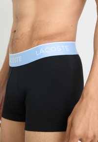 Black cotton boxer briefs with a light blue waistband featuring white "LACOSTE" text; smooth texture, fitted design.