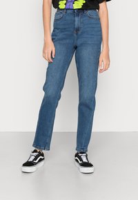 Jeans de denim azul de cintura alta com pernas retas, apresentando passantes para cinto e um leve efeito de desgaste. Combinados com sapatilhas pretas.