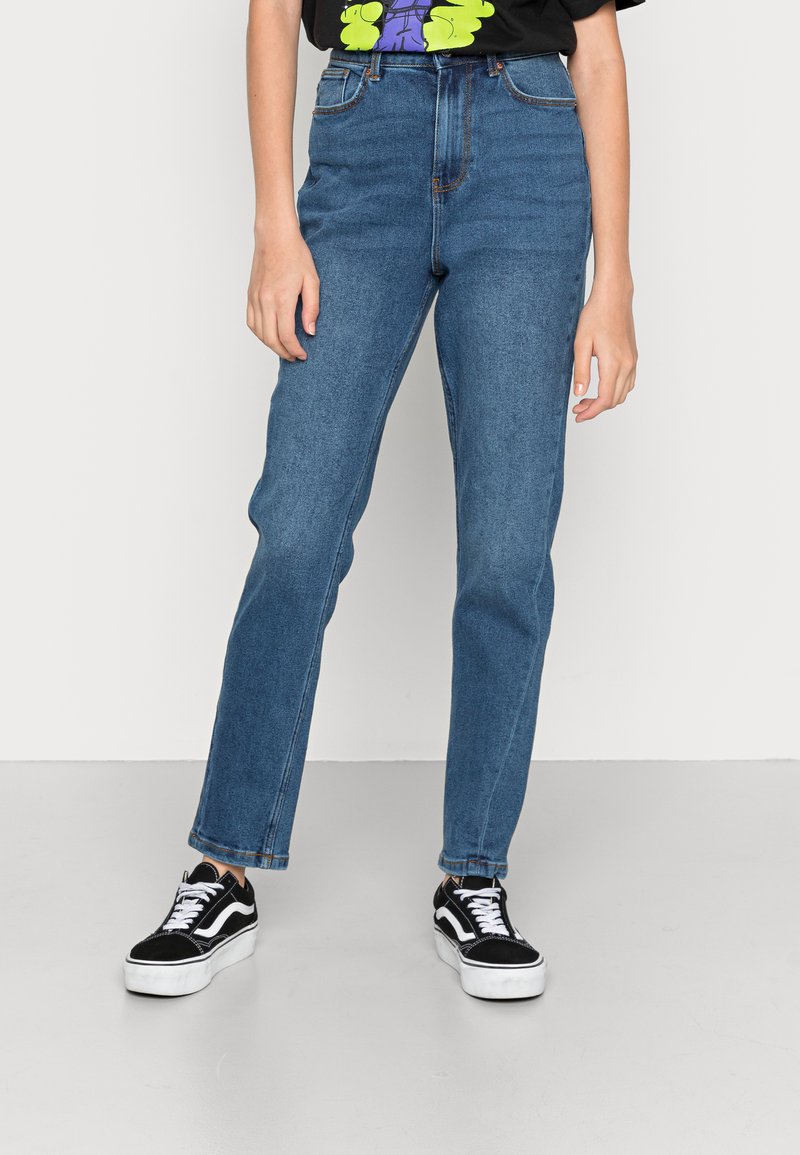 Jeans de denim azul de cintura alta com pernas retas, apresentando passantes para cinto e um leve efeito de desgaste. Combinados com sapatilhas pretas.