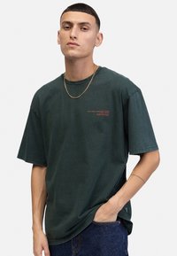 T-shirt en coton vert foncé à manches courtes, présentant un petit logo en texte rouge sur le devant et une coupe décontractée. Une chaîne en or est incluse.