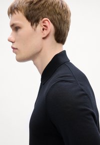 Μαύρο πόλο μπλουζάκι με ριγωτό γιακά. Η ομαλή υφή και η εφαρμοστή σχεδίαση αναδεικνύουν τη σιλουέτα του χρήστη. Αποτυπώνεται από το πλάι.