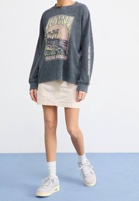 Sweat-shirt gris à manches longues avec un motif de plage, associé à une mini-jupe beige clair. Le modèle porte des chaussettes blanches aux chevilles et des baskets grises.