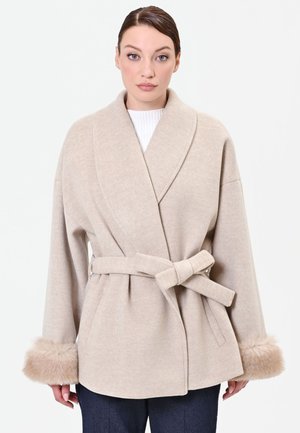 Donna che indossa un cappotto di lana beige con cintura, ampio colletto e polsini con rifiniture in pelliccia, abbinato a pantaloni scuri e top bianco.