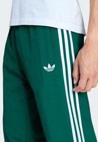 Zielone spodnie sportowe wykonane z lekkiego materiału, z białymi paskami i haftowanym logo na udzie. W zestawie elastyczny pasek w talii.