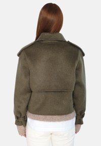 Oliven grønn ullblandingsjakke med høy krage, oppbrettede ermer og dobbeltspente design. Har et lagdelt bakdesignet for ekstra tekstur.