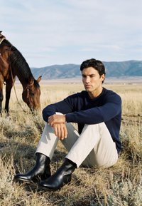 Homme en pull bleu marine et pantalon clair assis sur de l’herbe sèche dans un champ, un cheval broutant à proximité, avec des montagnes en arrière-plan.