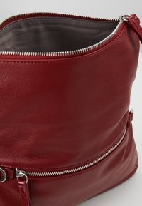 Červená kožená taška se splývavým designem, stříbrným zipem a šedou podšívkou. Obsahuje externí zipovou kapsu pro další úložný prostor.