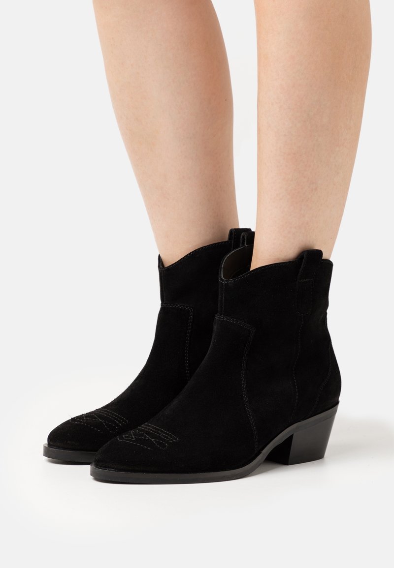 Jambes portant des bottines en daim noir à talons bas inclinés et coutures décoratives sur les orteils sur fond blanc.