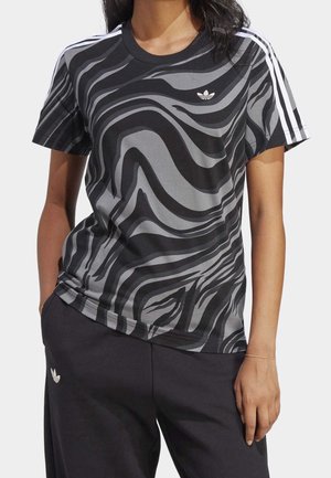 Kobieta ubrana w czarną i szarą koszulkę Adidas z wirującym wzorem, białe paski na rękawach oraz czarne spodnie z logo Adidas.