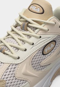 Μπεζ και εκρού αθλητικό παπούτσι FILA με επάνω μέρος από δίχτυ, χοντρά κορδόνια, λογότυπα FILA στη γλώσσα και στο πλάι, και επενδεδυμένο γιακά για άνεση.