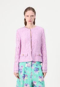 Spitzenjacke in hellem Lavendel mit runder Ausschnitt, Frontverschluss und zwei Taschenklappen mit goldenen Akzenten. Kombiniert mit floral gemusterten Hosen.