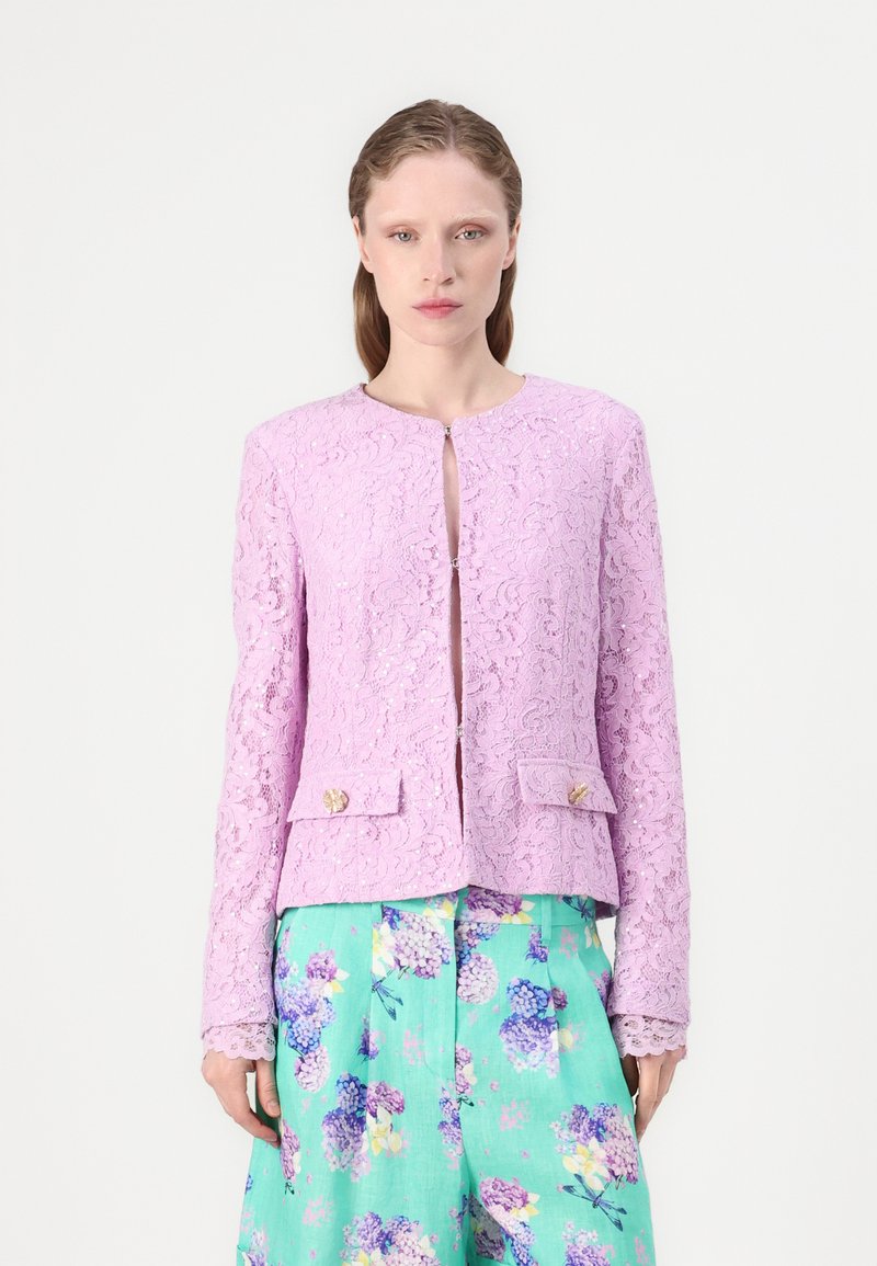 Spitzenjacke in hellem Lavendel mit runder Ausschnitt, Frontverschluss und zwei Taschenklappen mit goldenen Akzenten. Kombiniert mit floral gemusterten Hosen.