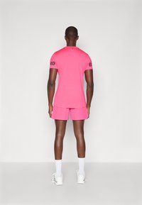 Rózsaszín sportpóló és rövidnadrág szett, rövid ujjú, fekete "BRG" felirattal az ujjakon. Fehér zoknival és tornacipővel viselve; sima szövetfelület.