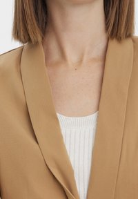 Blazer beige à texture lisse, avec un large revers et une coupe ajustée, superposé sur un haut blanc côtelé.